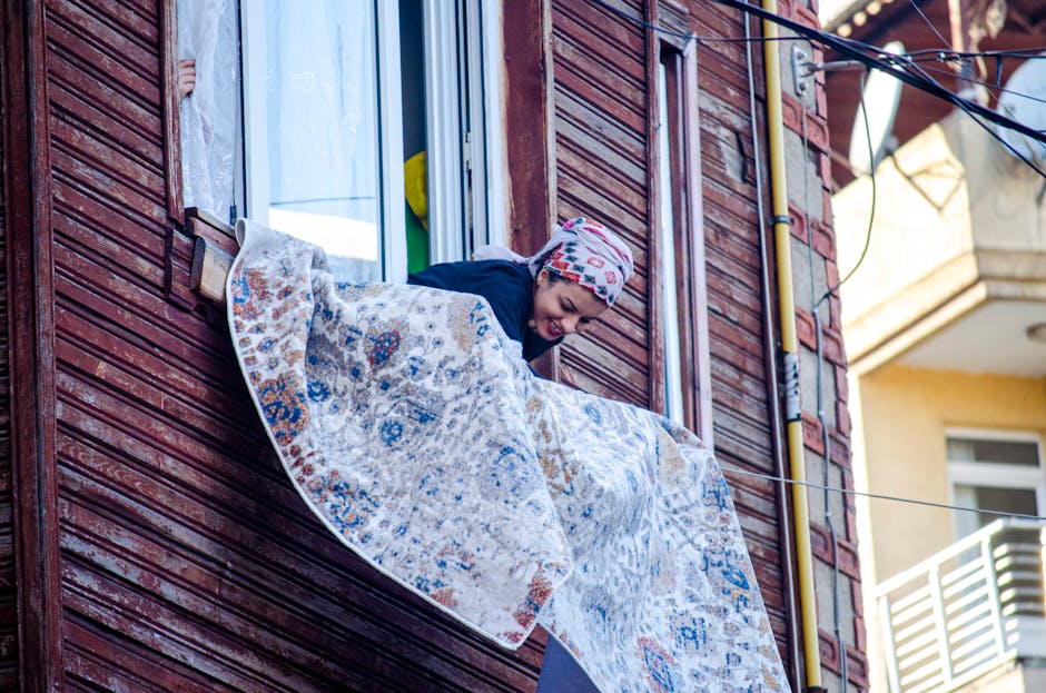 A person leaning out of a window of a wooden building, hanging laundry on a clothesline. The individual is wearing a colorful patterned headscarf and a dark jacket, with a cheerful expression. The bui