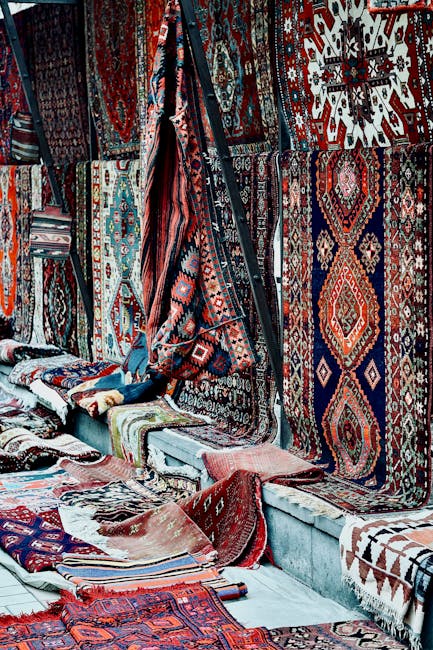 A person leaning out of a window of a wooden building, hanging laundry on a clothesline. The individual is wearing a colorful patterned headscarf and a dark jacket, with a cheerful expression. The bui